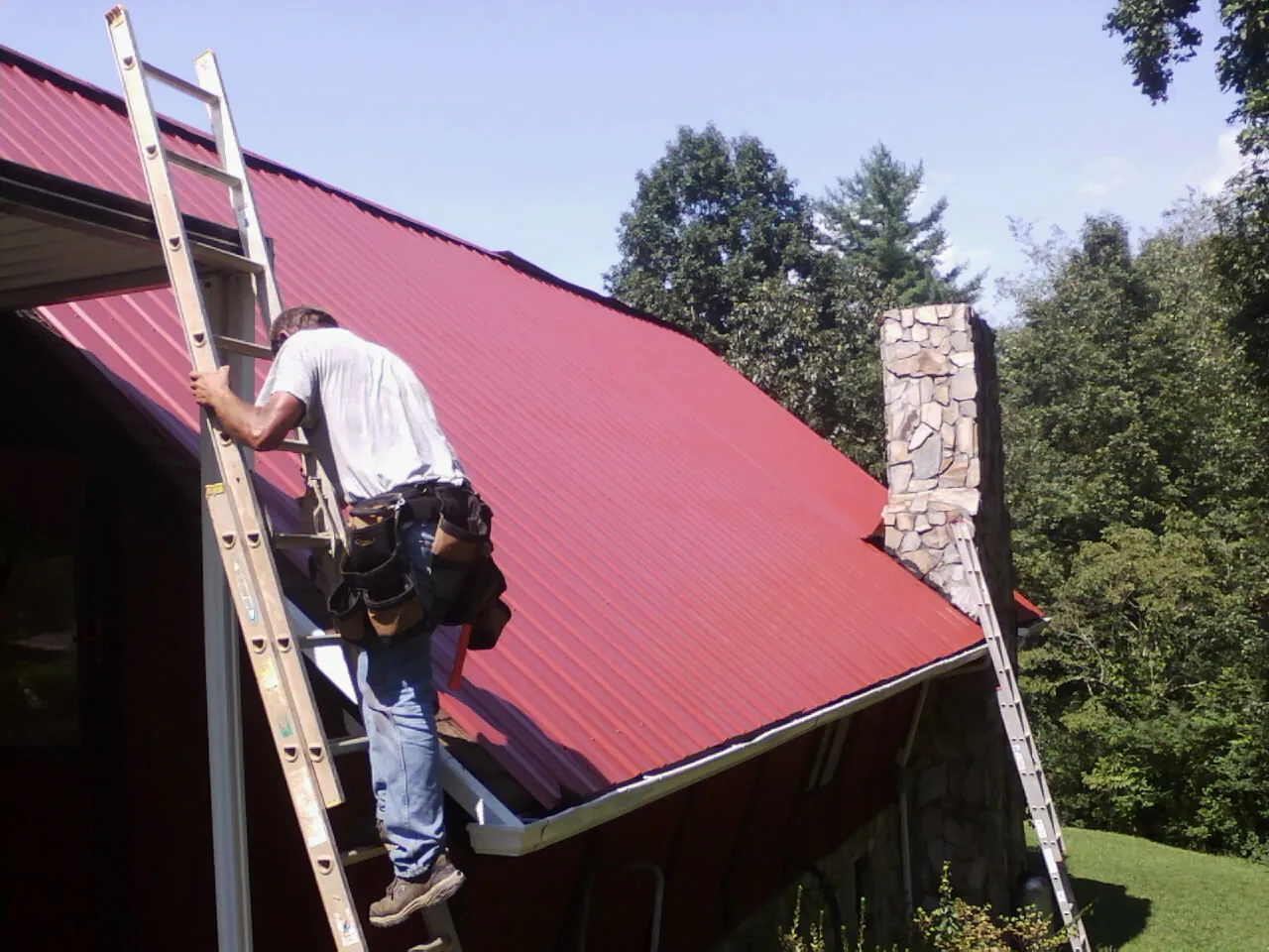 Roofer on a ladder beside a red metal roof and stone chimney — Soffit & Fascia Repair by Roofing Brothers in Sunnyside